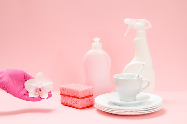 Woman's hand in a nitrile protective glove with an orchid flower, bottles of dishwashing liquid, white plates, a saucer, a cup on the pink background. Washing and cleaning concept.