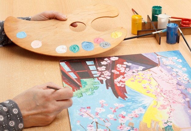 Женская рука рисует японский пейзаж в помещении на деревянном столе с палитрой