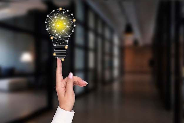 A woman's hand points to a glowing light bulb on the background of the office. Concept of innovation, ideas in business. High quality photo