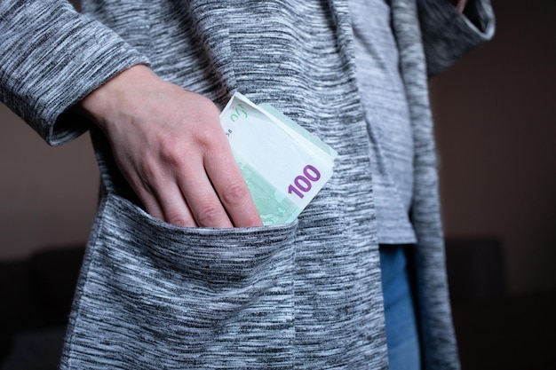 Woman's hand puts or takes out paper money 100 euro into the pocket of a gray jacket cardigan