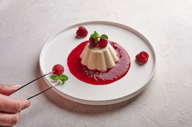 Woman's hand puts tweezers leaf mint to panna cotta with syrup, raspberries and mint