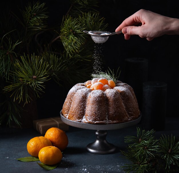 La mano di una donna spruzza zucchero a velo su una torta di natale al mandarino. immagine scura e d'atmosfera