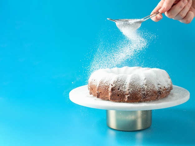 Photo woman's hand sprinkling icing sugar over fresh home made bundt cake powder sugar falls on fresh perfect bunt cake over blue background copy space for text ideas and recipes for breakfast or dessert