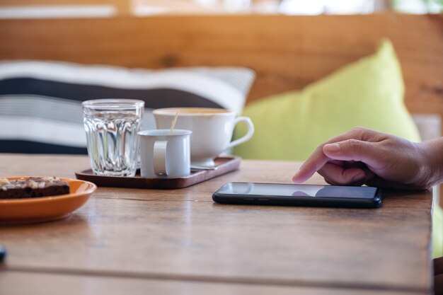 カフェの木製テーブルにコーヒーカップとスマートフォンの画面を使用して触れている女性の手