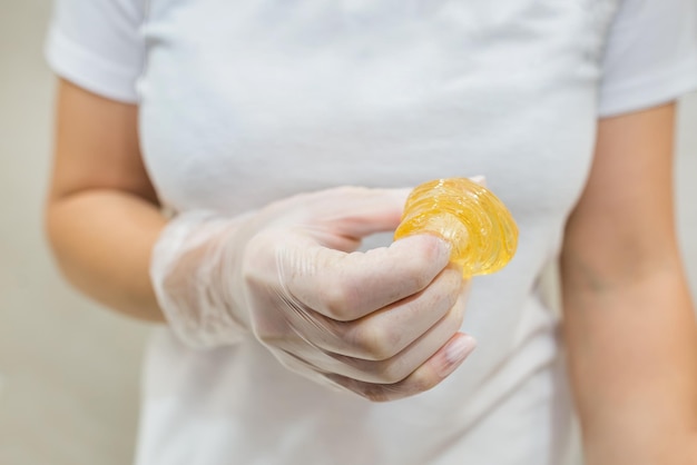 A woman's hand in a white glove holds a yellow sugar paste for depilation Epilation depilation Rolled ball of sugar paste in beautician's hand