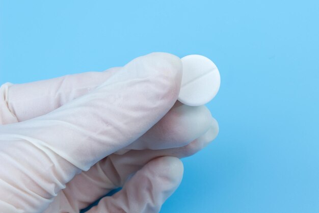 Женская рука в белой медицинской перчатке держит таблетку крупным планом на синем фоне. Концепция профилактики вирусных заболеваний.