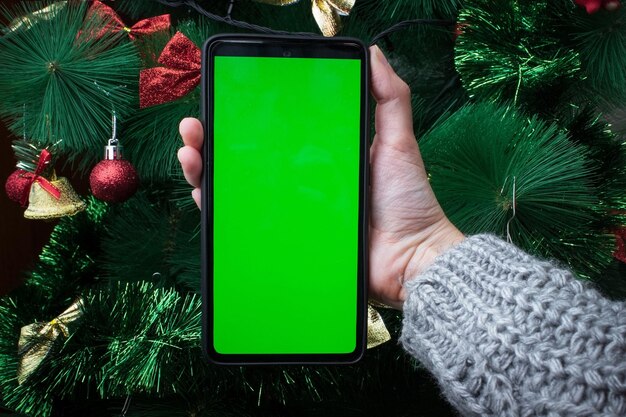 겨울 스웨터를 입은 여성의 손이 장식된 크리스마스 트리 배경에 수직 위치에 녹색 화면이 있는 스마트폰을 들고 있습니다