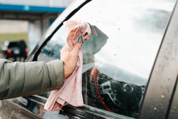 Женская рука вытирает тряпкой мокрое чистое окно автомобиля после мытья автомобиля, вид сбоку крупным планом