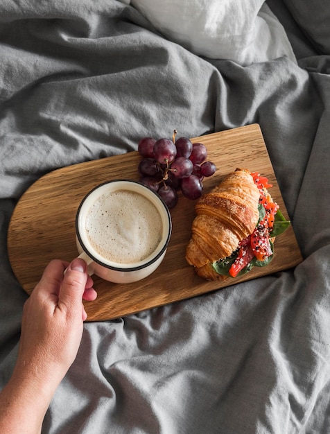 Женская рука с чашкой капучино и сэндвичем с круассаном с лососем на доске в разобранной кровати. Медленное уютное утро выходного дня в постели.
