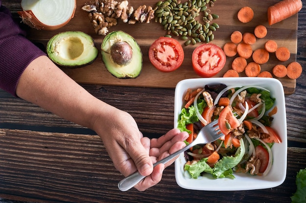 Woman's hand with fork piercing the vegetables of a Vegetable salad on vintage wooden background. Healthy vegetarian food, top view.