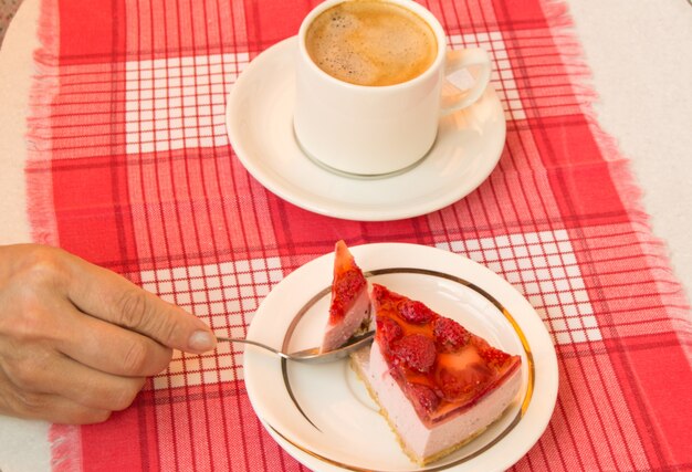 A woman's hand with pleasure holds a piece of fresh strawberry cheesecake, a cup of coffee on a saucer, a view from above. The concept of a delicious gourmet breakfast.