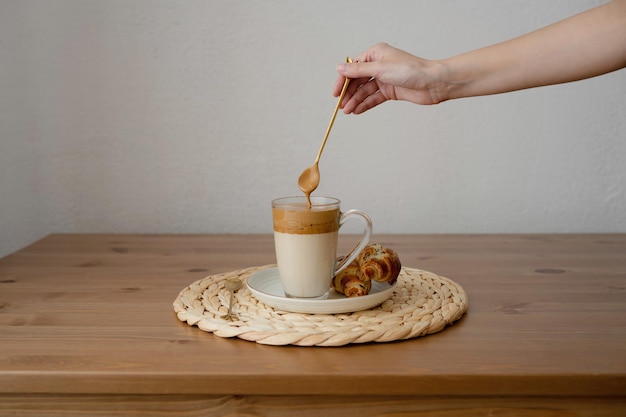 A woman's hand with a spoon over a coffee cup with milk and croissants on a wooden table