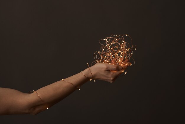 A woman's hand wrapped in a Christmas garland holds a fire in her hand on a dark background