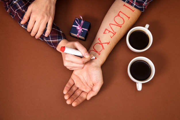 Woman's hand writes red letters i love you on man's hand near two cup of coffee on brown.