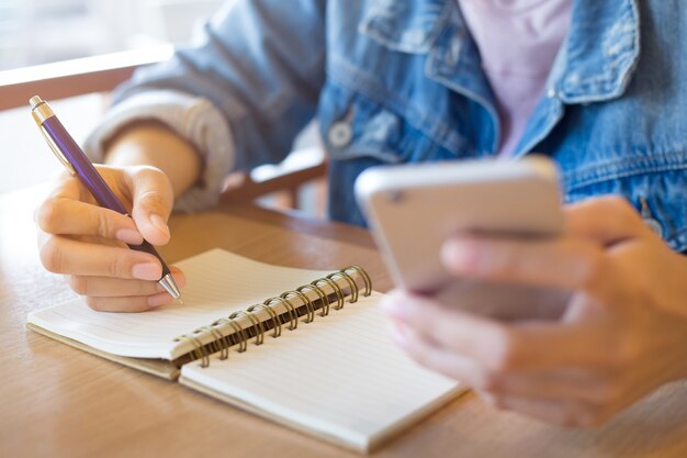 Woman's hand writes spiral sheet and holds the phone on a wooden desk.