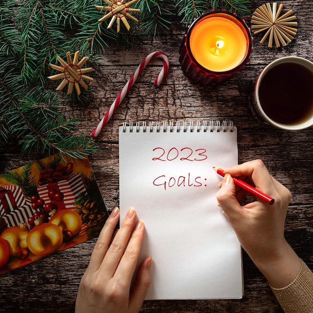 Woman\'s hand writing 2023 goals on a blank notepad sheet. dark\
wooden background, christmas decor