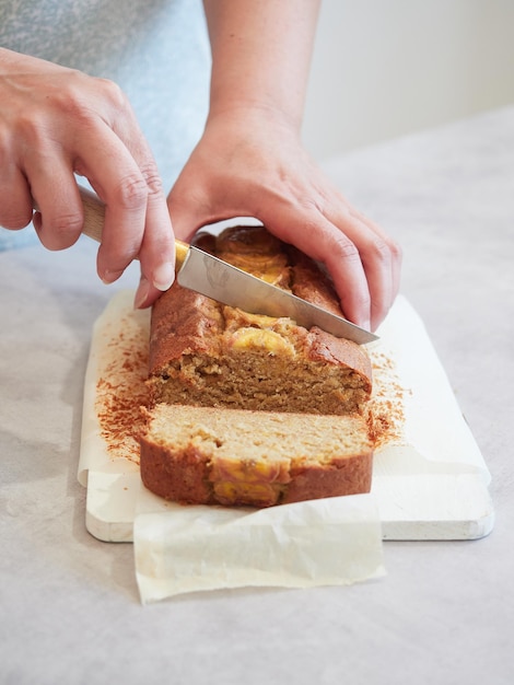 Woman's hands cutting freshly baked banana bread on white cutting board with baking paper on the table