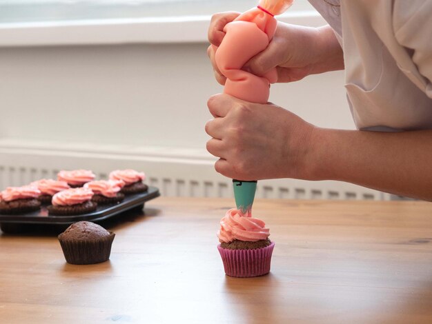 The woman's hands hold a pastry bag and squeeze out pink cream on cupcakes making cakes