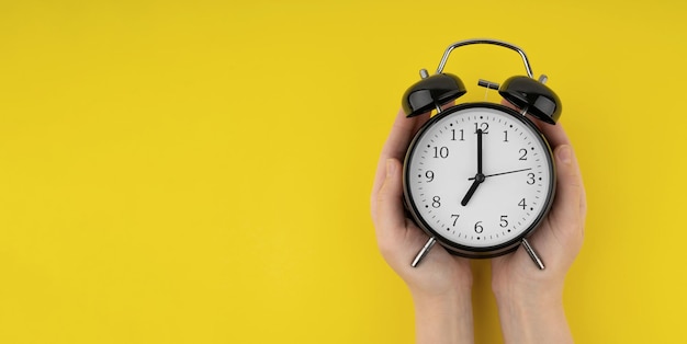 Woman's hands holding a black alarm clock on a yellow background