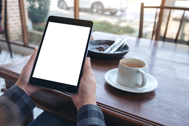 woman's hands holding black tablet pc with blank screen with coffee cup on wooden table in cafe