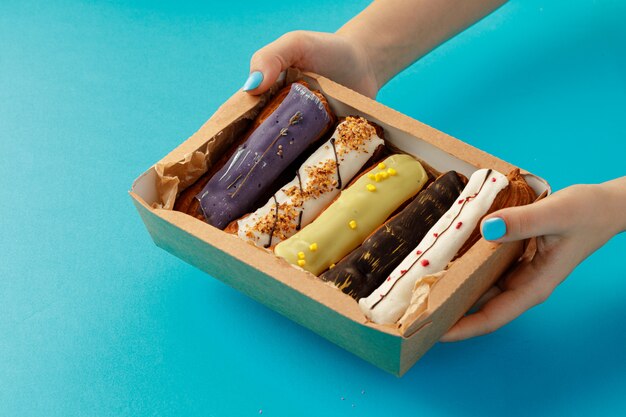 Woman's hands holding a box with fresh eclairs on blue background