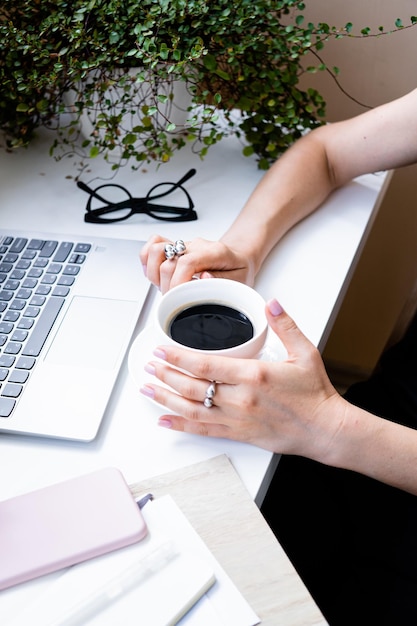 사진 노트북과 녹색 식물이 있는 아늑한 사무실에서 커피 한 잔을 들고 있는 여성의 손