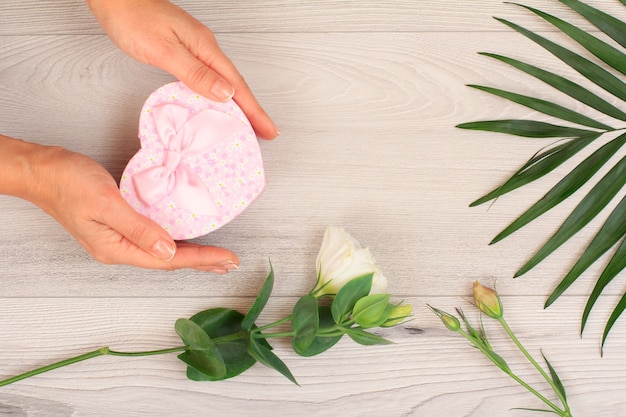 Woman's hands holding a gift box in heart shape over gray wooden background with beautiful flowers and green leaves. Concept of giving a gift on holidays. Top view.