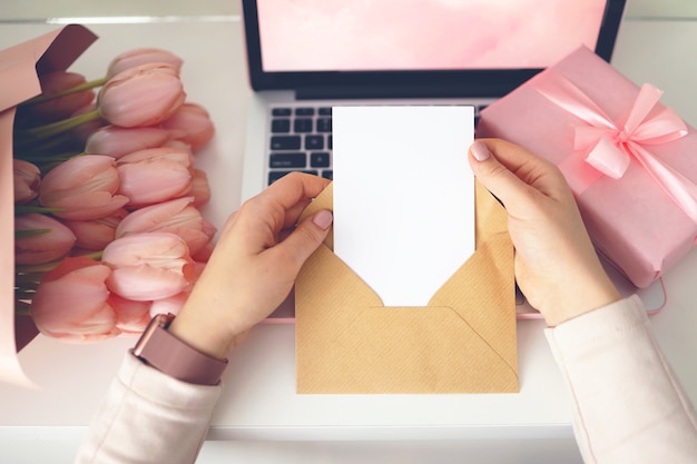 クラフト封筒に手紙を持っている女性の手。ピンクの背景、バレンタインデーのコンセプト。背景にチューリップの花とピンクのギフトボックス。レディースホームデスク。