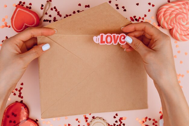 Woman's hands holding a letter in craft envelope on the rose background.  And puts the word love in an envelope. Valentine's day concept.