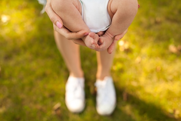 Woman's hands holding little baby in white clothing in her hands on the green grass