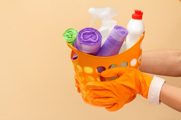 Woman's hands in rubber protective gloves holding orange basket with garbage bags, bottles of glass and tile cleaner, sponge on beige background. Washing and cleaning concept.