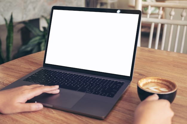 woman's hands typing and touching on laptop touchpad with blank white desktop screen while drinking coffee on wooden table in cafe