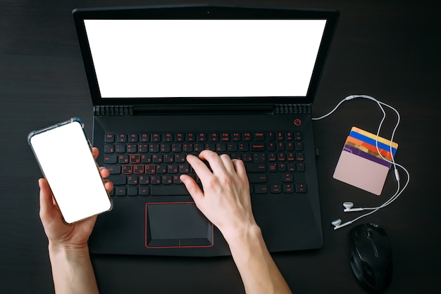 Woman's hands using laptop keyboard and smartphone for shopping online while holiday sales. Top view of the workspace with credit cards on black background. Blank screen for advertising.