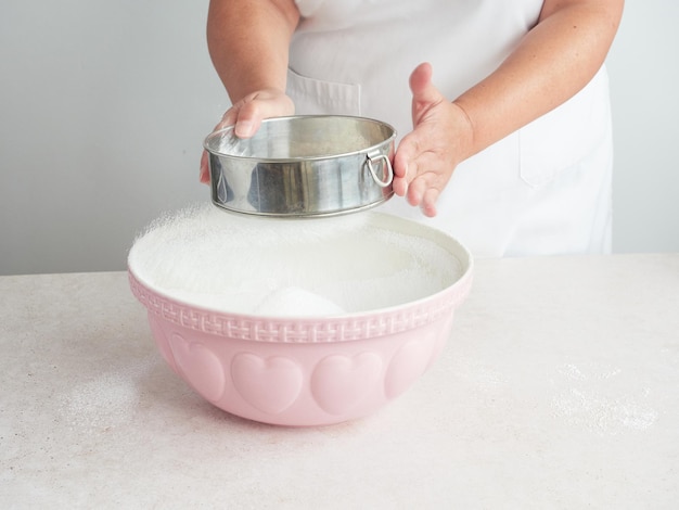 흰색 앞치마를 입은 여성의 손이 탁자 위의 커다란 분홍색 세라믹 그릇 위에 금속 체로 설탕을 체로 치고 있다