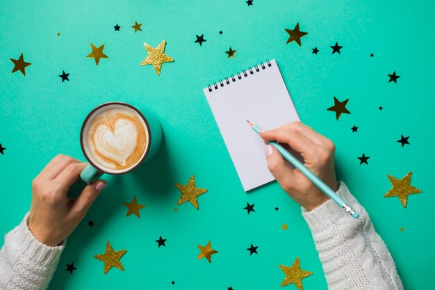 Woman's hands in white sweater making plans for next year writing them into notebook Making wishlist or New Year's resolutions concept Top view blue background with colorful stars Copy space