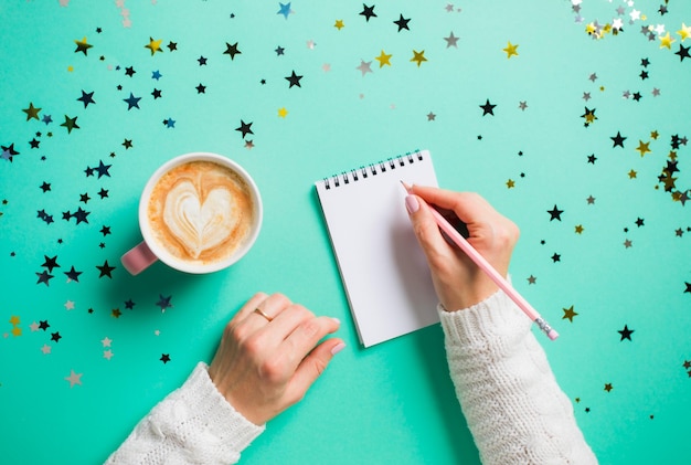 Woman's hands in white sweater making plans for next year writing them into notebook Making wishlist or New Year's resolutions concept Top view blue background with colorful stars Copy space