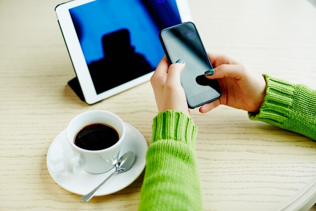 Woman's hands with dark manicure holding mobile phone, tablet and cup of coffee on table, freelance concept, online shopping, flat lay