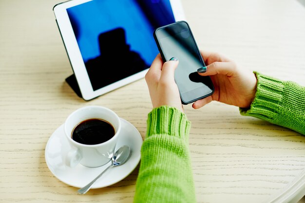 Woman's hands with dark manicure holding mobile phone, tablet and cup of coffee on table, freelance concept, online shopping, flat lay.