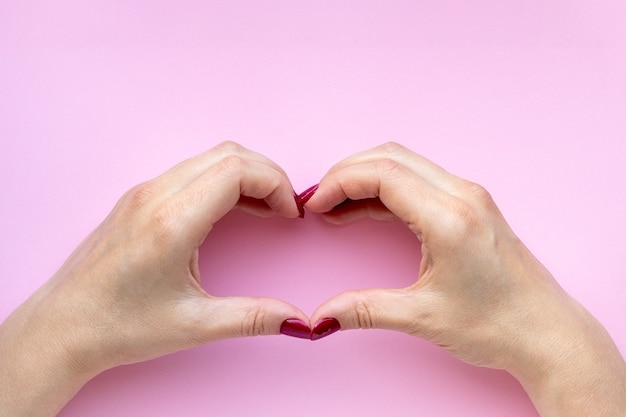 Foto mani della donna con le unghie di colore rosso che fanno il simbolo del cuore sulla parete rosa