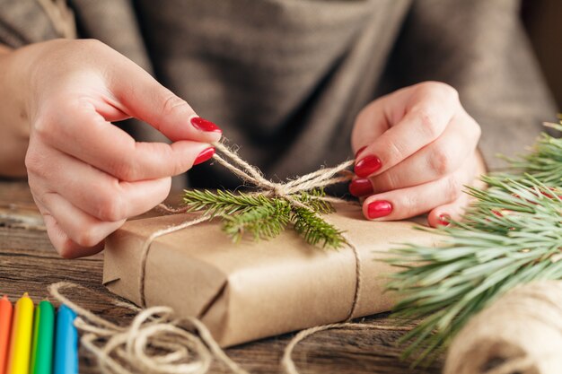 クリスマスの休日を包む女性の手は、ひものリボン付きペーパークラフトに存在します。雪の結晶で飾られたクリスマスギフトボックスで弓を作ります。白い木製のテーブルにはさみ