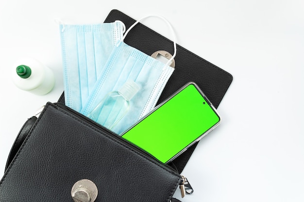 Woman's purse with phone, antiseptic, medical mask, and against white background, phone with green screen, chromakey, top view, isolated