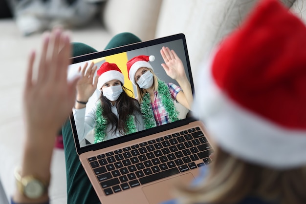 Woman in santa claus hat holds laptop on knees and communicates with her friends in protective masks