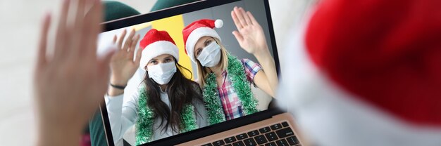 Woman in santa claus hat holds laptop on knees and communicates with her friends in protective masks