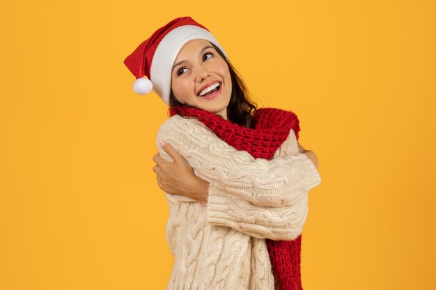 Woman in santa hat embracing herself on yellow backdrop