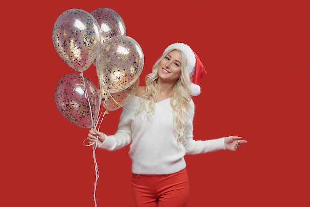 Woman in Santa hat holding balloons on a red wall. Christmas