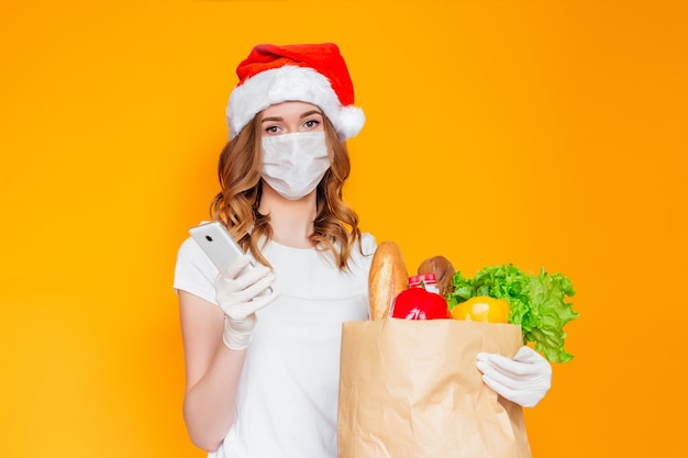 Woman in santa hat holds a mobile phone, a paper bag with food, vegetables, fruits, pepper, yogurt, salad, herbs, bread