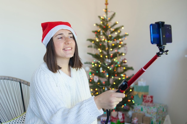 Женщина в новогодней шапке возле елки разговаривает по видеосвязи по телефону