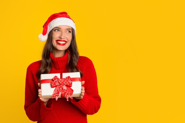 Woman in santa hat presents xmas gift against yellow backdrop