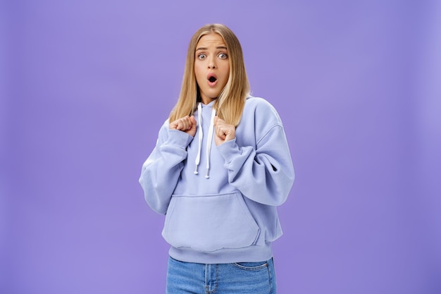 Woman scared of sudden loud noise in dark gasping looking shocked and concerned tilting backwards with frightened look pressing clenched fists to body from fear, wearing warm hoodie over purple wall