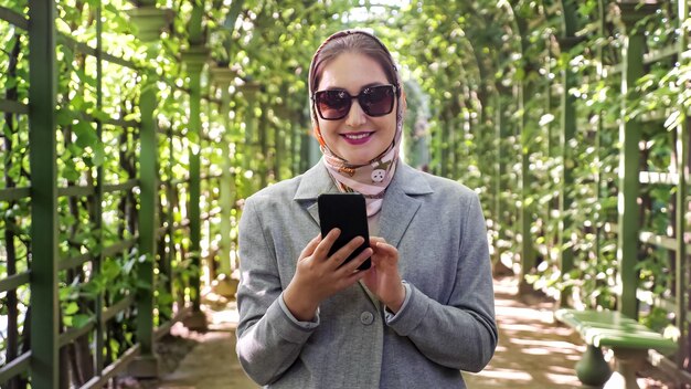 Woman in a scarf, jacket and sunglasses typing text on the phone in the garden arch.